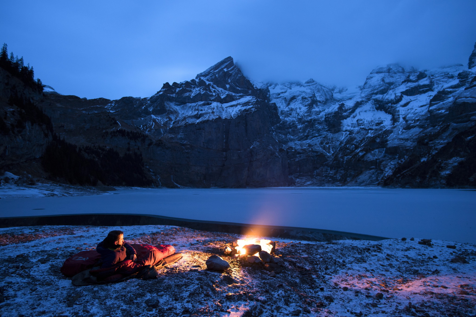 Mężczyzna w śpiworze patrzy na ognisko naprzeciw zamarzniętego ogniska Oeschinensee, Szwajcaria.