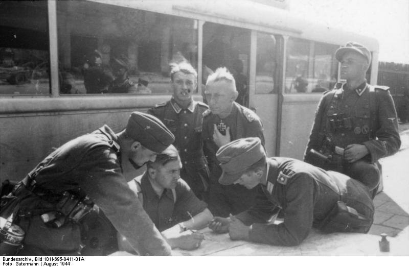 SS-Gruppenführer Heinz Reinefarth (po środku z Krzyżem Rycerskim) podczas walk na Woli, dowodził z autobusu sztabowego ustawionego przy skrzyżowaniu ul. Syreny i Wolskiej. Drugi z lewej to przypuszczalnie informator, który przedostał się z terenów powstańczych i wskazuje na mapie polskie pozycje, sierpień 1944 r. (fot. Gutermann, Bundesarchiv, Bild 101I-695-0411-01A, CC-BY-SA).